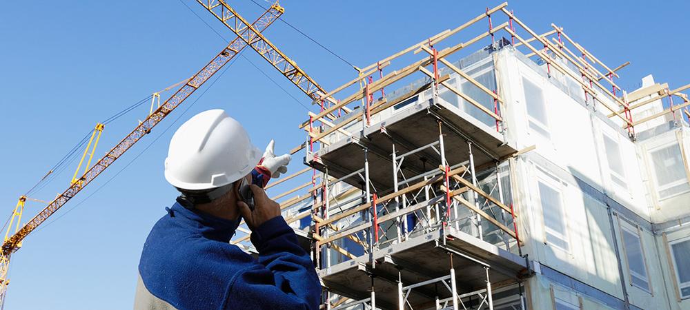 A man wearing a hardhat chats on a phone and gestures toward a building under construction.