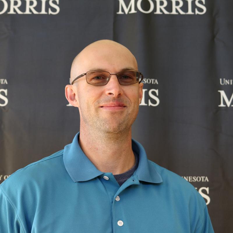 A bald man wearing shaded glasses, wearing a teal polo shirt and standing in front of a black backdrop with the words, University of Minnesota Morris 