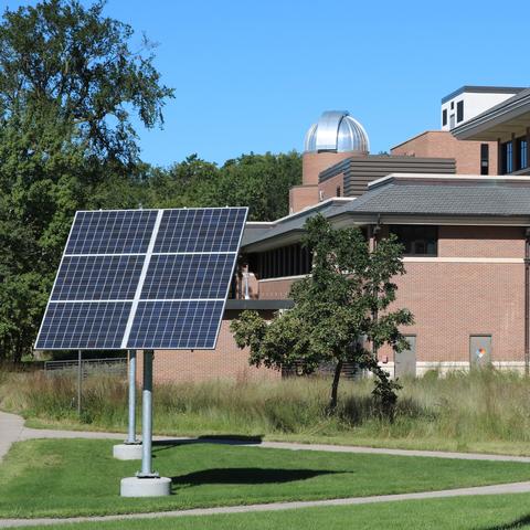 Two large raised solar panels on poles, in front of a brick building with trees in the landscape