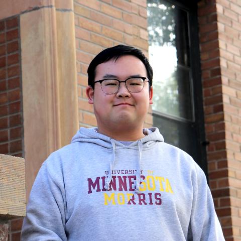 A young Chinese man standing outside of a brick building.  He has short black hair, and black-rimmed glasses and is wearing a grey sweatshirt with the words, University of Minnesota Morris on it. 