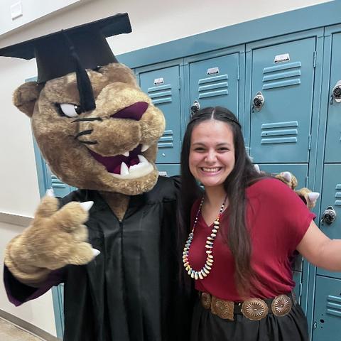 The UMN Morris Cougar mascot in a cap and gown with Kianna Big Crow, an Indigenous woman with long dark hair awearing a red shirt.  